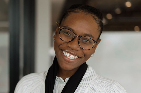 Latrina wearing a white blouse wiht a black scarf around her neck, wearing glasses and smiling into the camera