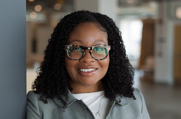 Markeia Scruggs has dark curly hair and glasses. She wears a grey collared jacket and smiles at the camera.